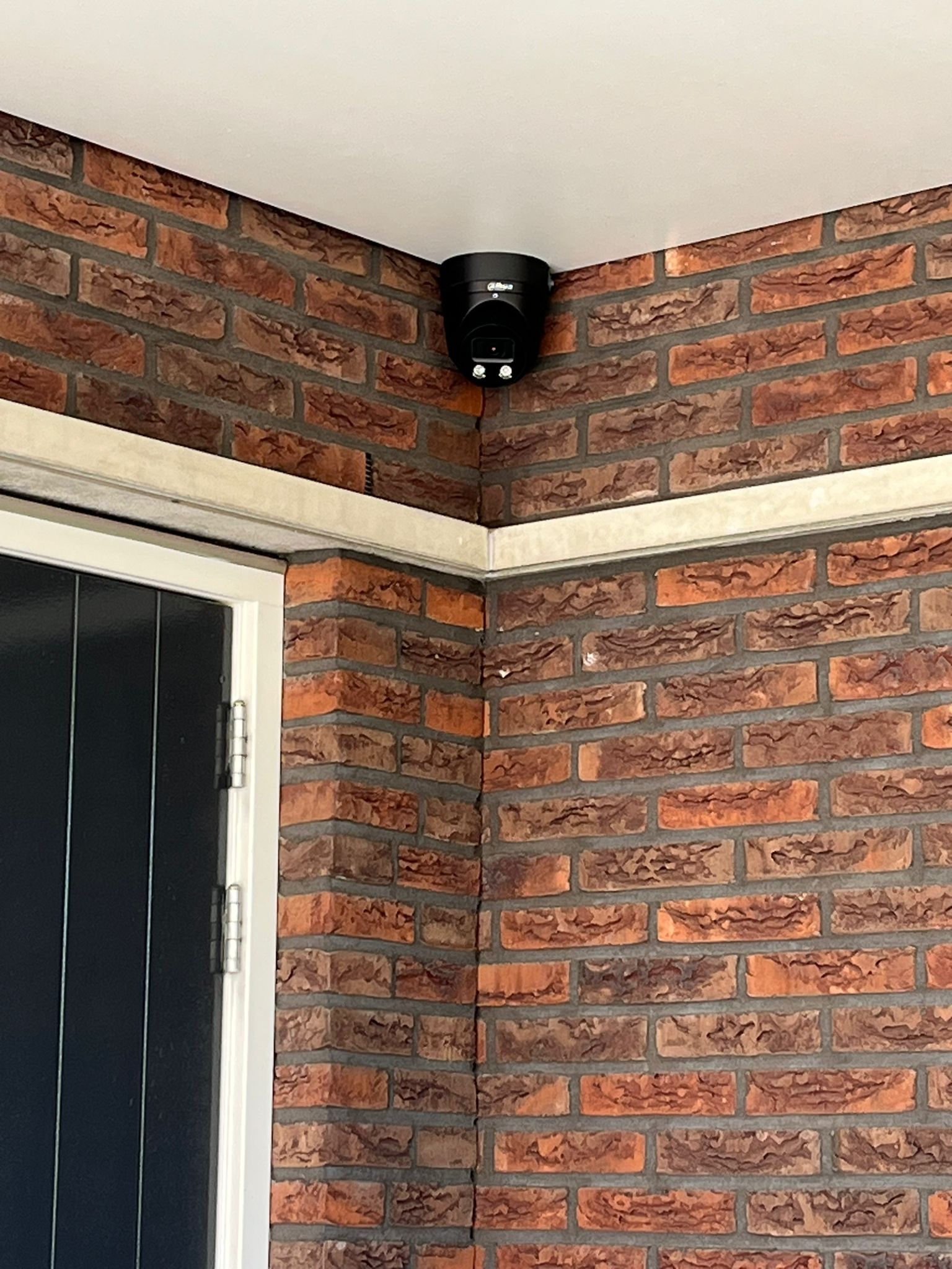 Zwarte Dahua turret-camera met Dahua-logo, netjes in de hoek gemonteerd onder een wit plafond in een overdekte entree met rode bakstenen muren