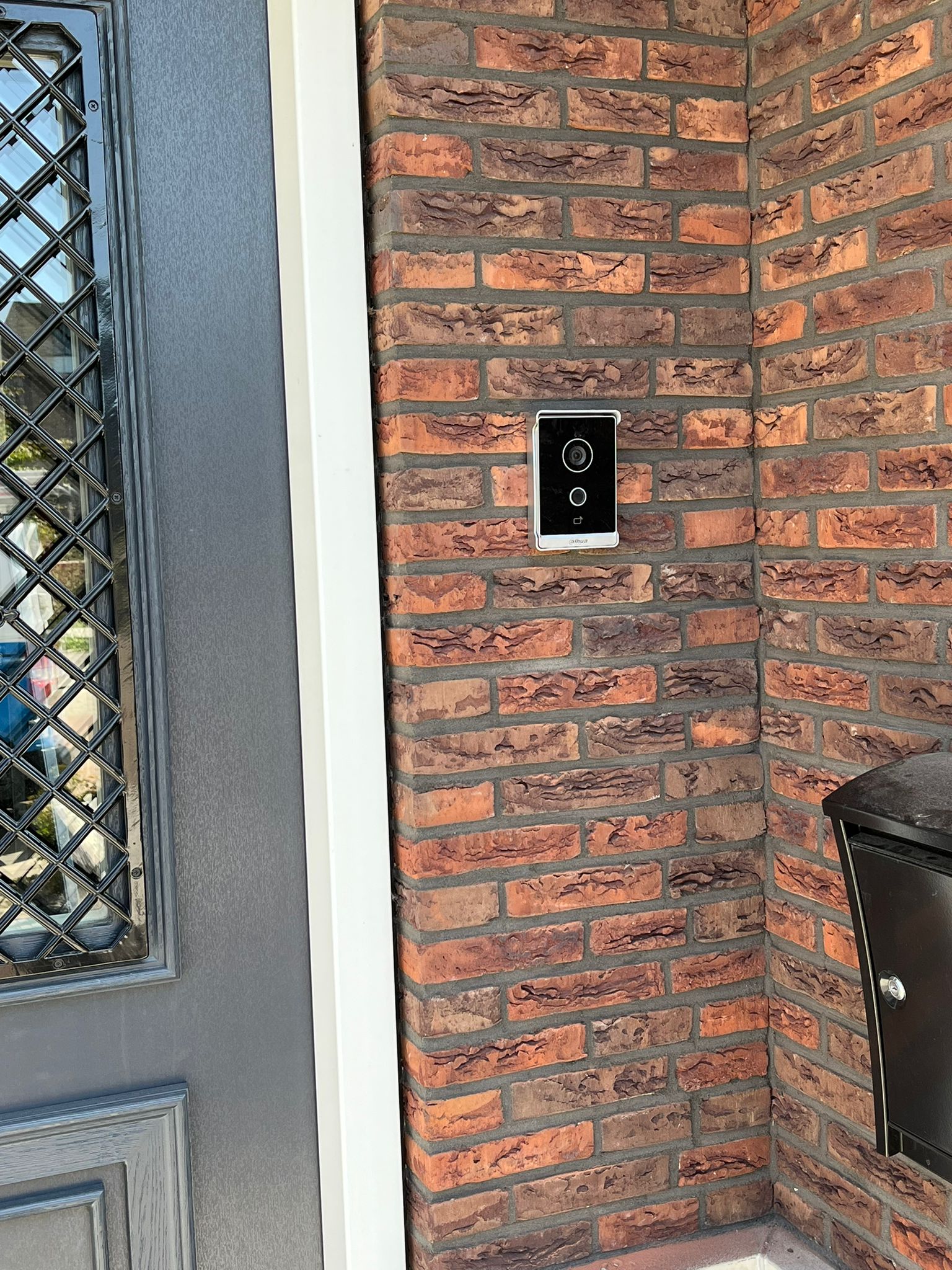 Dahua videodeurbel met camera netjes gemonteerd op de rode bakstenen muur naast een grijze voordeur met glas-in-lood