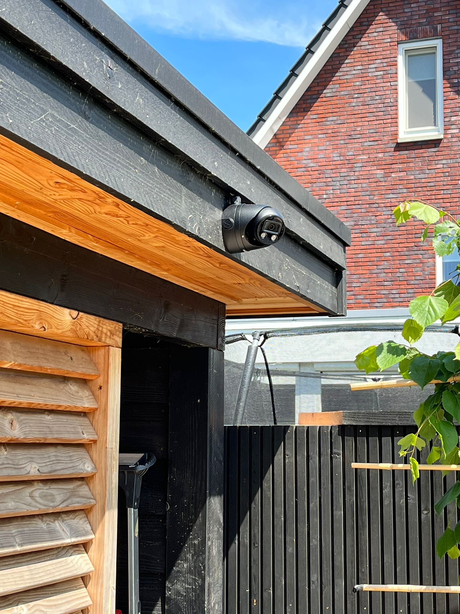 Zwarte Dahua camera onder de dakrand van een tuinberging, met de bakstenen gevel en het raam van de woning op de achtergrond in de zon