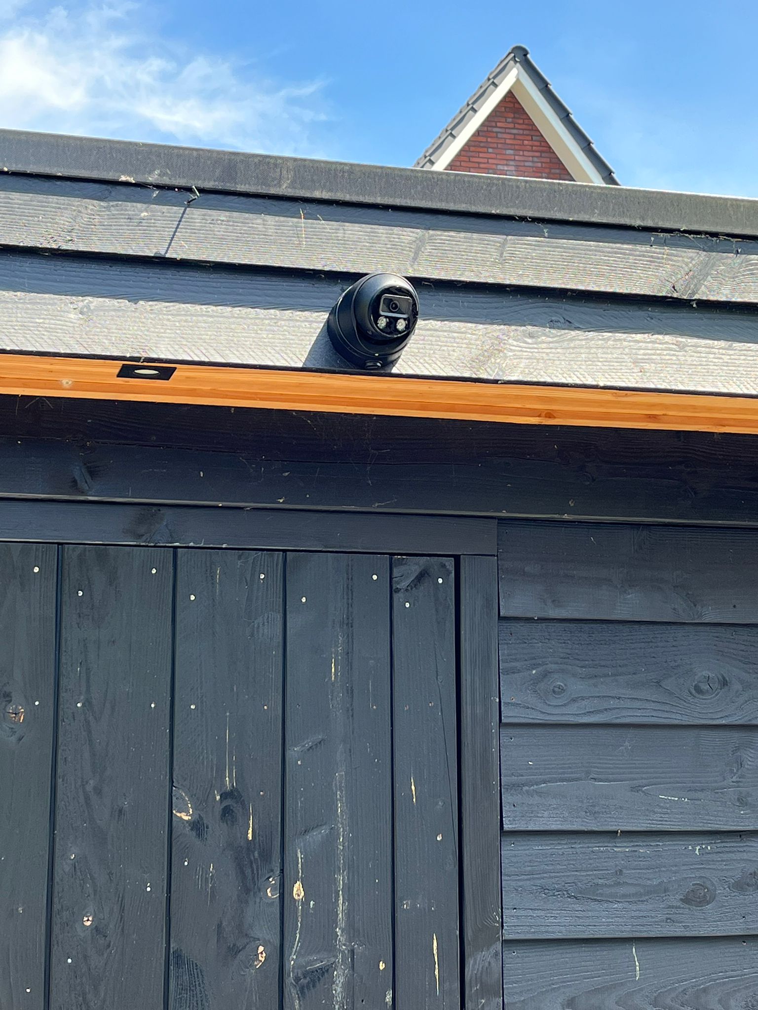 Zwarte Dahua turret-camera strak gemonteerd onder de dakrand van een donker houten bijgebouw, met de bakstenen woning op de achtergrond
