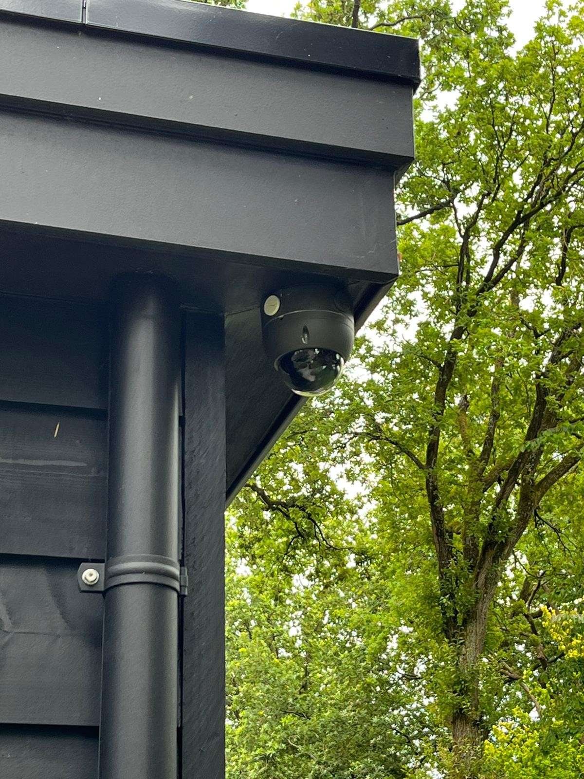 Zwarte Dahua dome-camera gemonteerd onder de zwarte dakrand van een gebouw, met een eik op de achtergrond