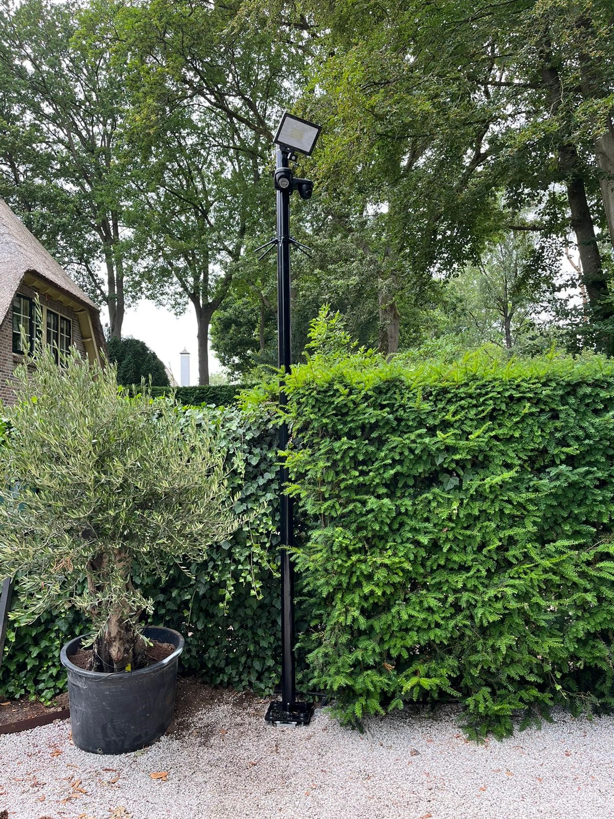 Zwarte camerapaal met camera en schijnwerper naast een groene haag, met een olijfboom in een pot en een rietgedekte woning op de achtergrond