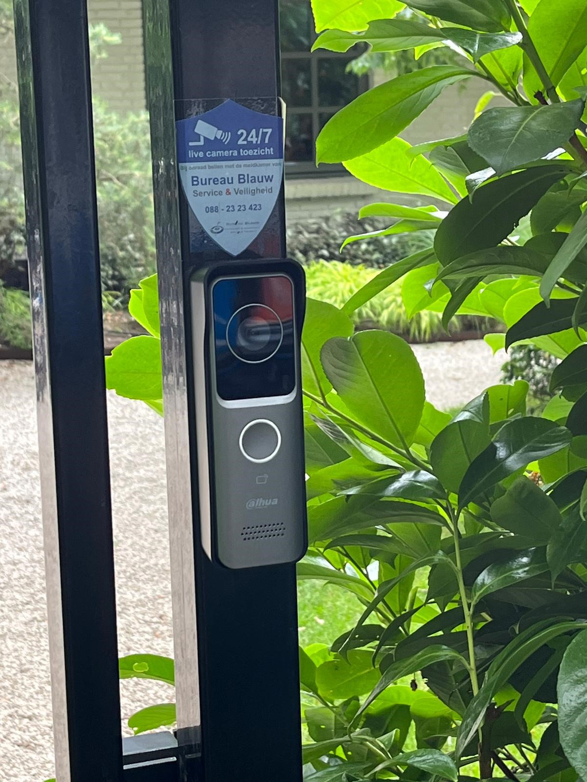 Dahua videodeurbel met camera en belknop gemonteerd op een zwarte toegangspoort, met daarboven een sticker met 24/7 cameratoezicht