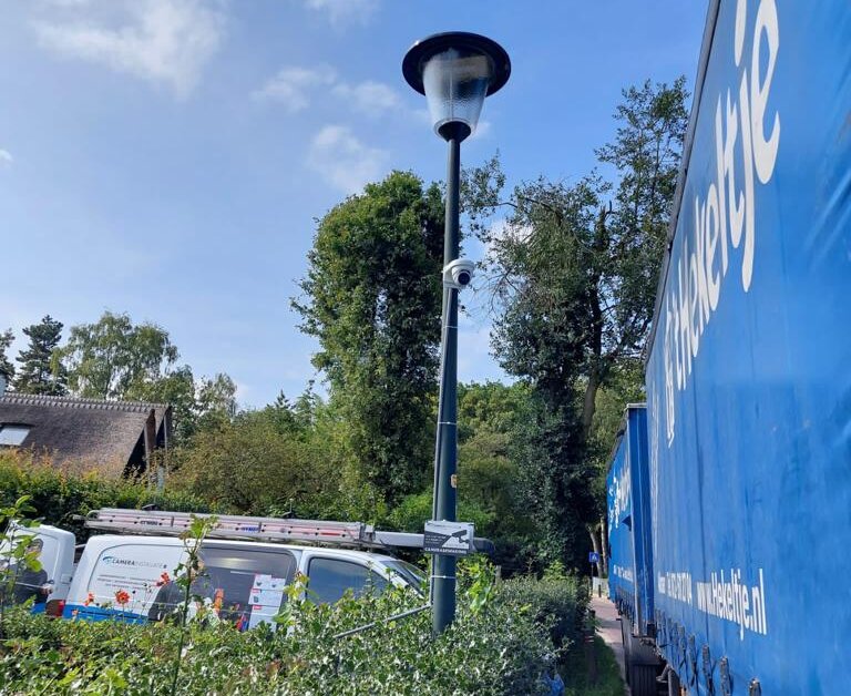 Camera gemonteerd op een lantaarnpaal langs een weg, met een servicebus van CameraInstallatie ernaast