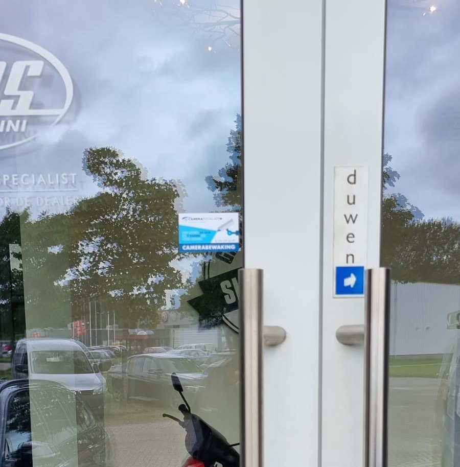 Glazen entreedeur van Garage Roos met een camerabewaking-sticker van CameraInstallatie, met geparkeerde auto's weerspiegeld in het glas