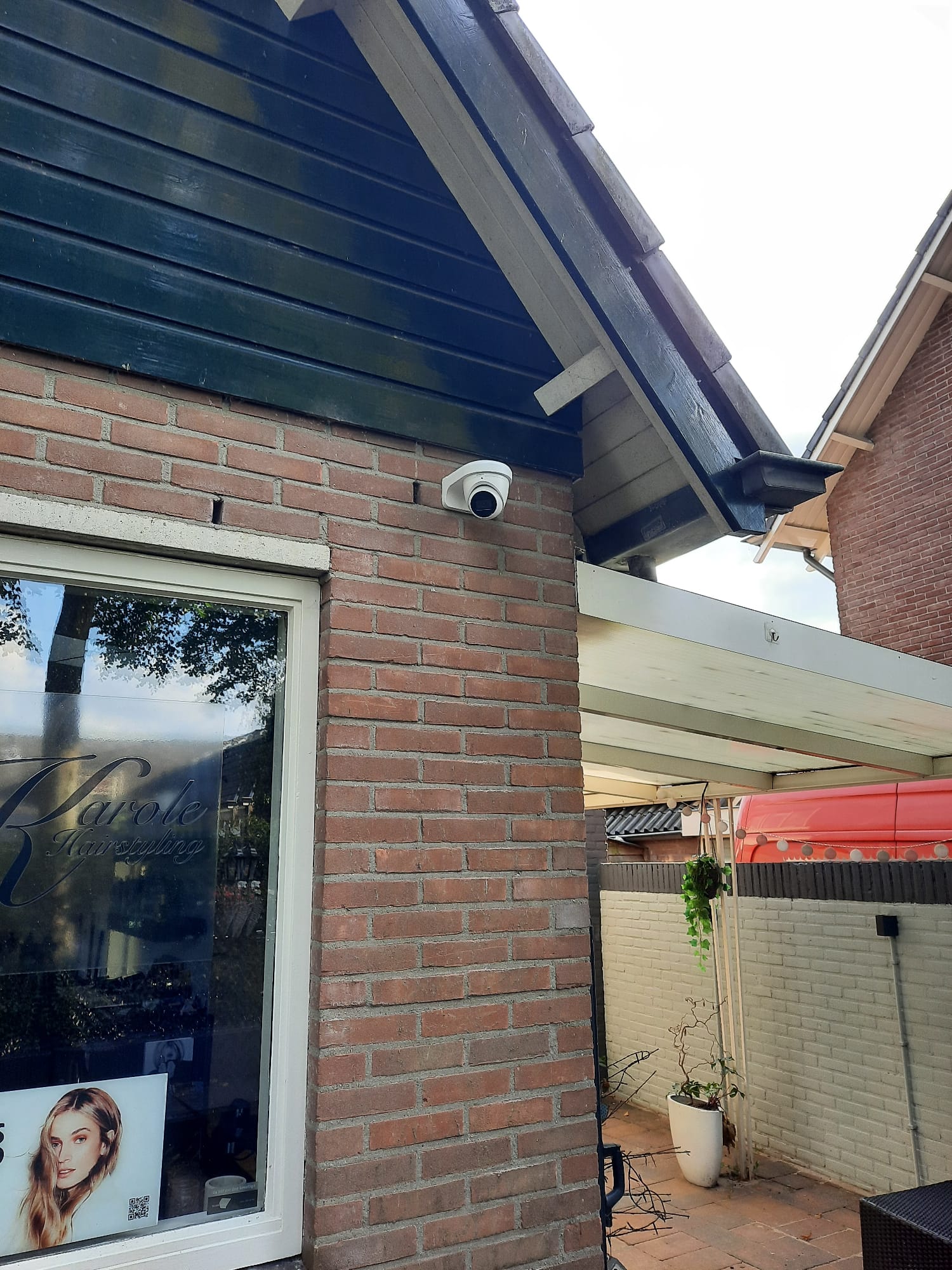 Witte dome-camera gemonteerd op de bakstenen hoek van een woning, onder de donkere houten daklijst