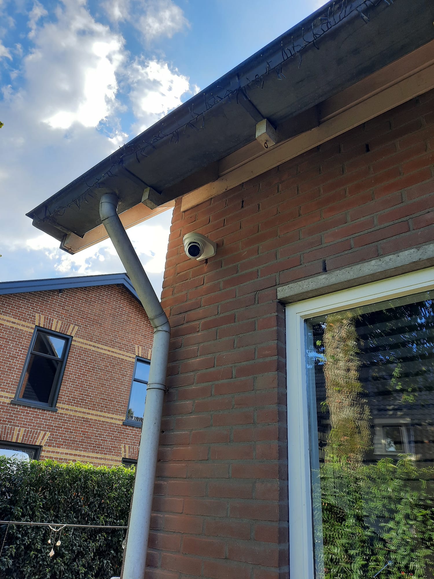 Witte dome-camera op de bakstenen hoek van een woning tegen een blauwe lucht, met een naastgelegen huis op de achtergrond