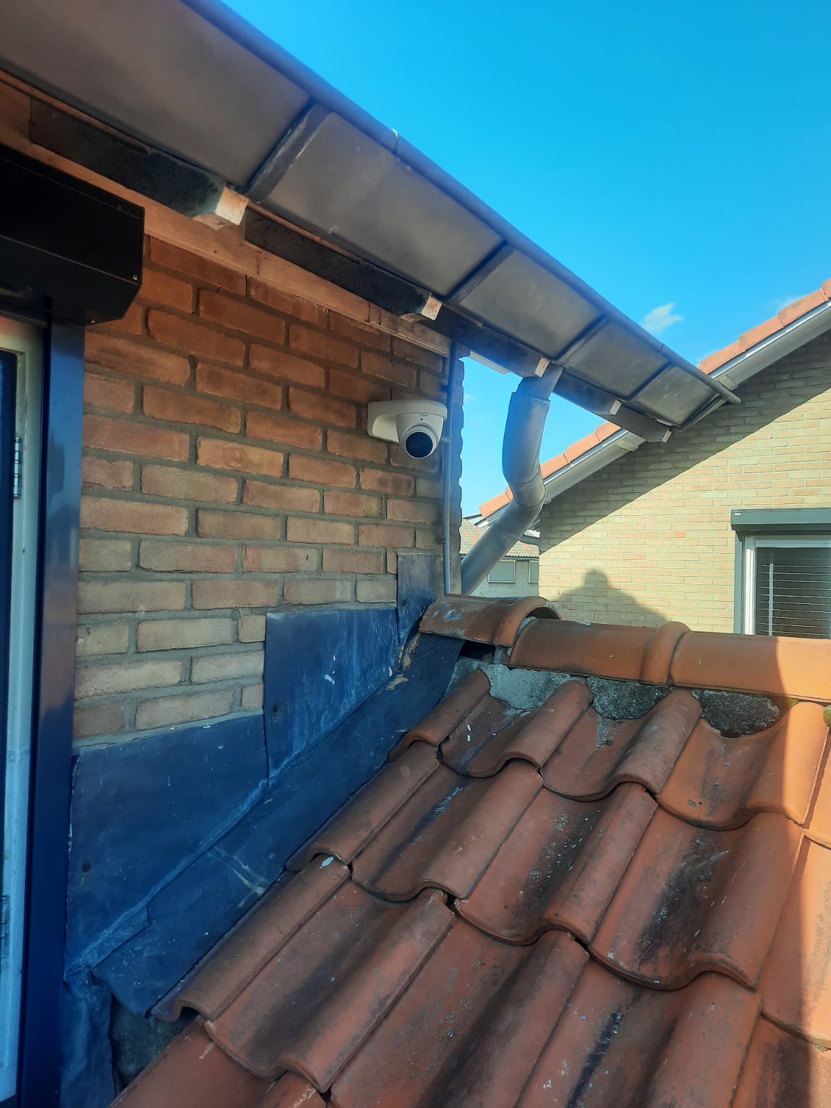Witte dome-camera vlak onder de dakrand van een woning, met dakpannen en een strakblauwe lucht