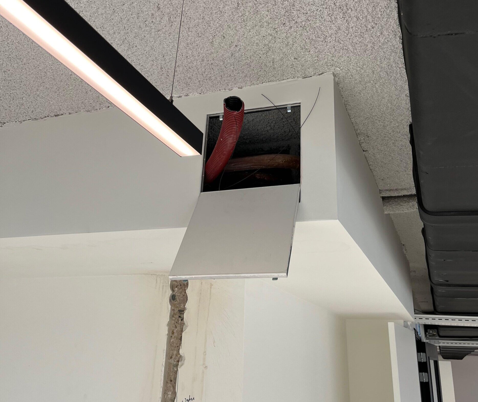 Openstaand inspectieluik in een verlaagd plafond met een rode kabelbuis en bekabeling die naar de verdieping erboven loopt