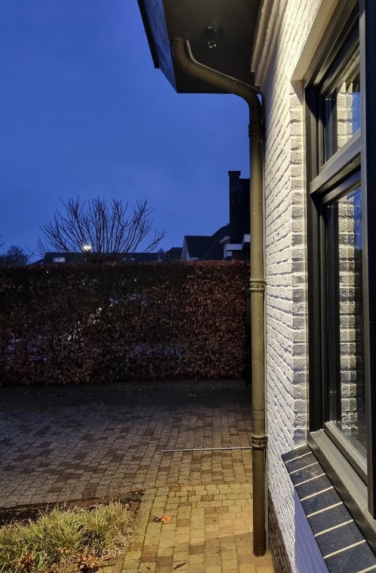 Schemerfoto langs de zijgevel van de woning; onder de dakrand is de camera zichtbaar, de tuin en de haag liggen in het donker