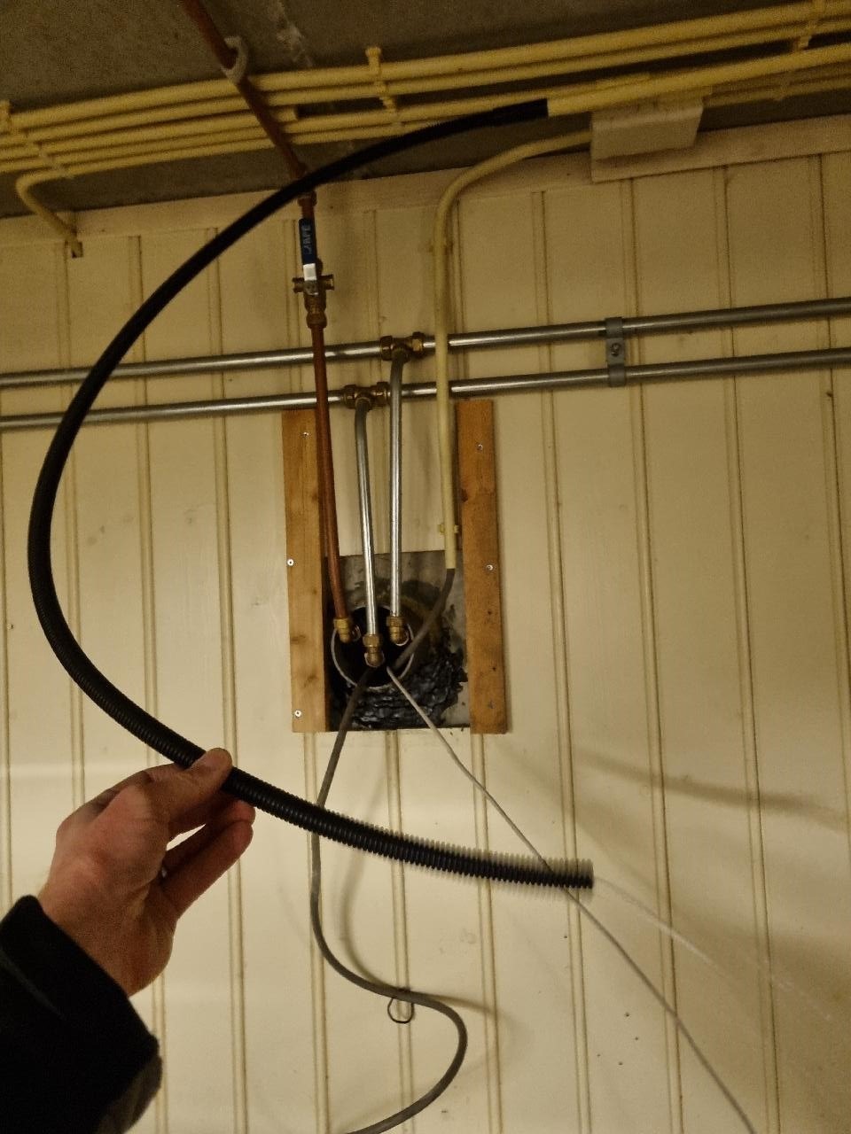 Monteur houdt een zwarte flexibele buis vast bij een muurdoorvoer met koperen leidingen in een kruipruimte