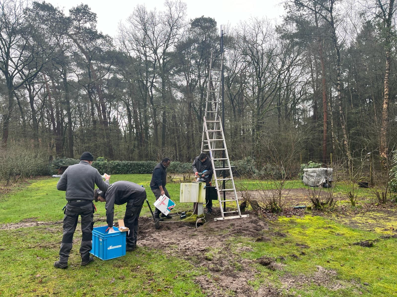 Vier monteurs plaatsen samen een mast op het grasveld, met een ladder tegen de mast en materiaal in een kist