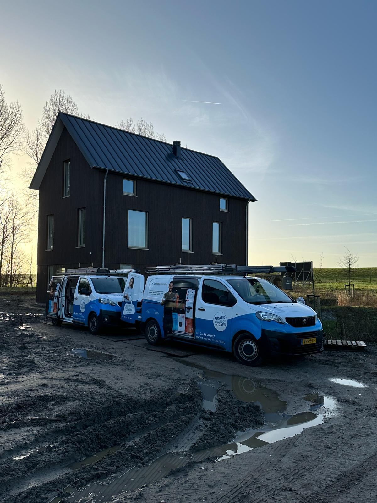 Twee bussen van CameraInstallatie staan op het nog onverharde bouwterrein voor de donker afgewerkte nieuwbouwwoning