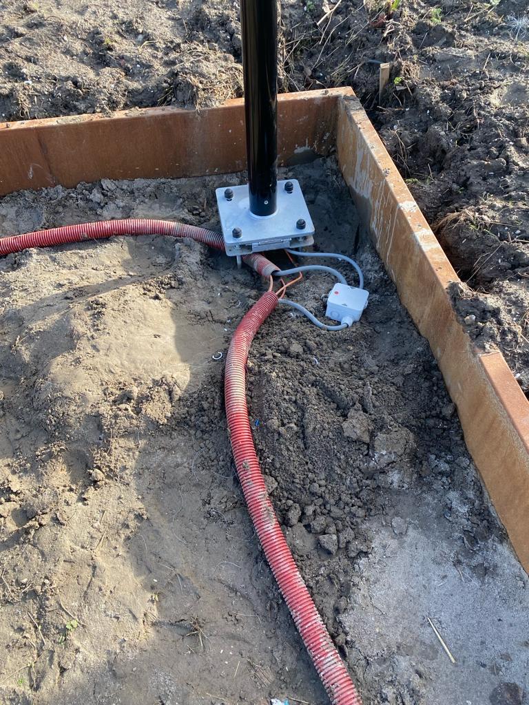 Voetplaat van een mast op een verse betonfundering, met rode mantelbuizen die van onderaf de mast in komen