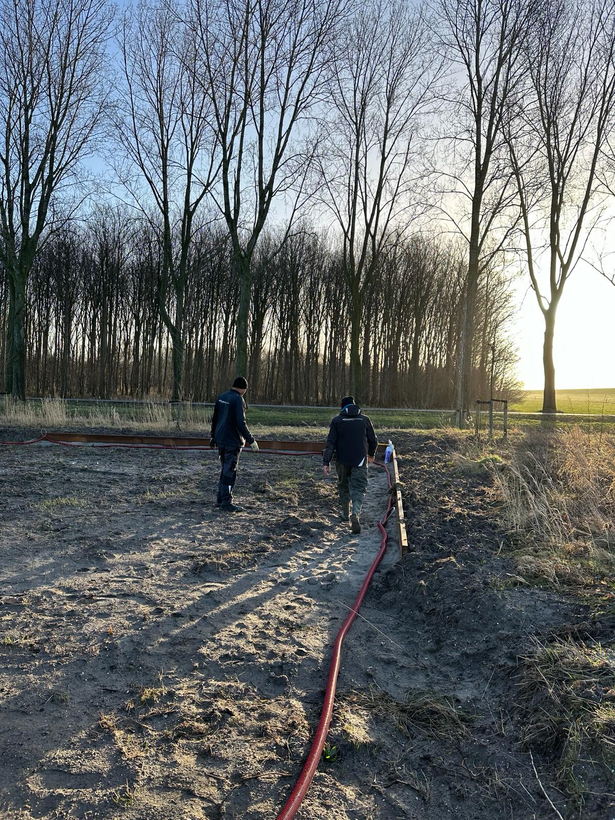 Twee monteurs lopen langs de open sleuf met rode mantelbuis over het terrein, met bomen op de achtergrond