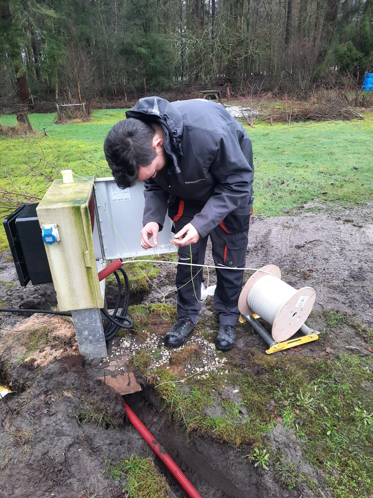 Monteur van CameraInstallatie werkt bij een groene aansluitkast op het terrein, met een kabelhaspel ernaast en een rode mantelbuis in de sleuf