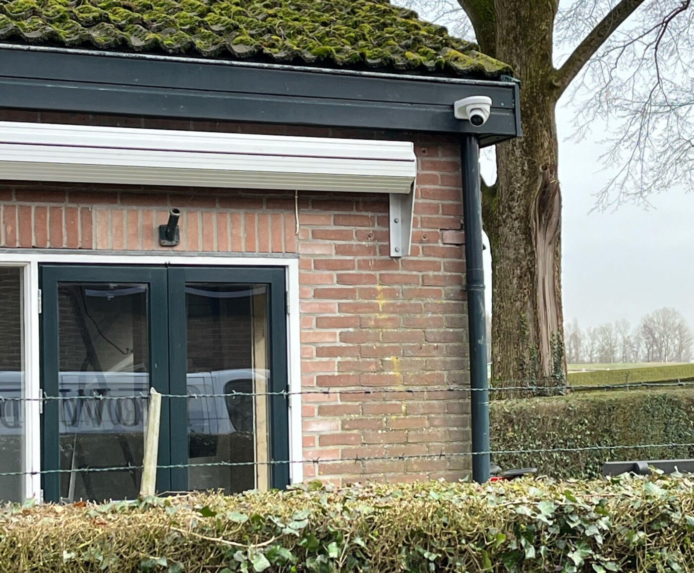 Witte dome-camera onder de dakrand van een bakstenen gebouwtje met uitzicht over het aangrenzende veld