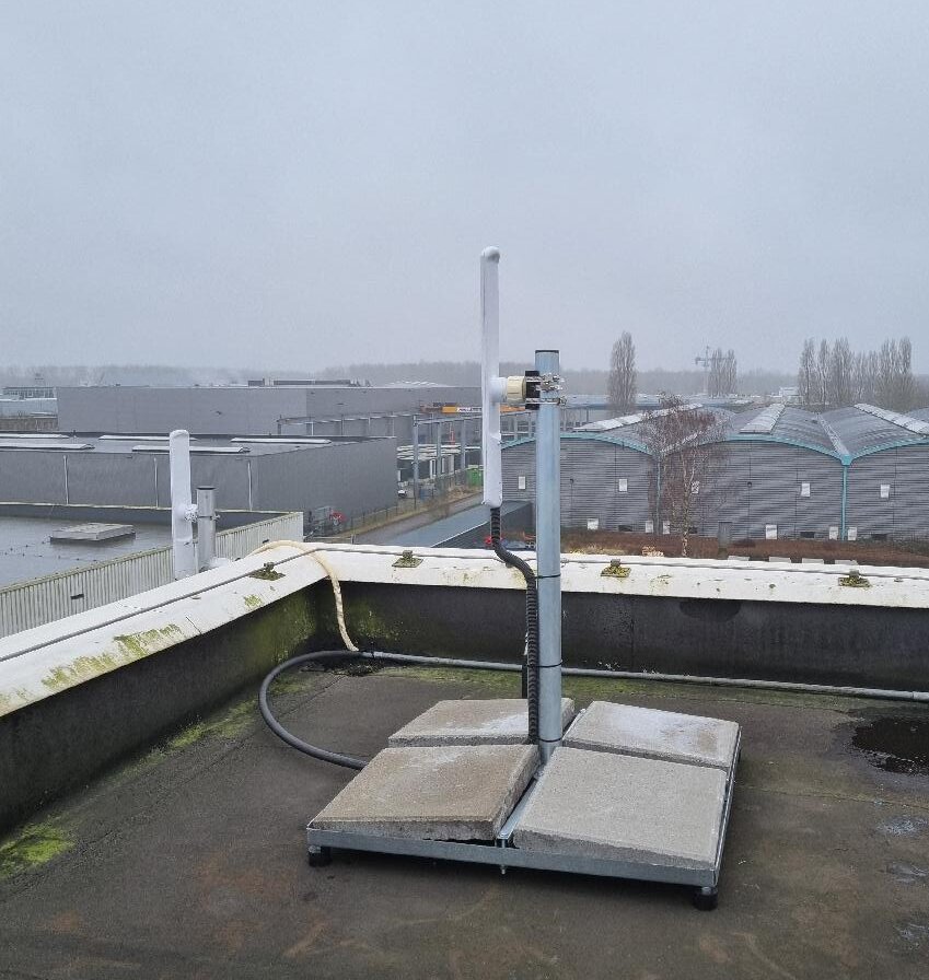 Straalzender op een verzwaarde voet op een plat dak, met de kabel die naar binnen loopt en een bedrijventerrein op de achtergrond