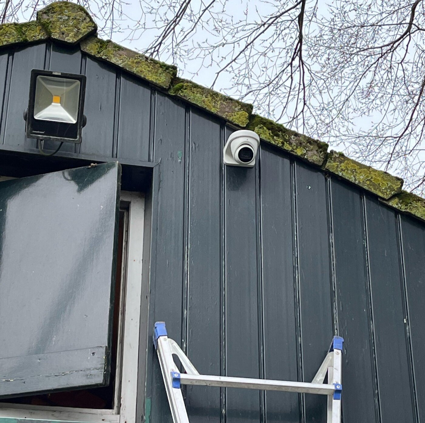 Witte dome-camera gemonteerd op een donkerblauw houten schuurtje onder de dakrand, met een ladder ertegenaan tijdens de installatie