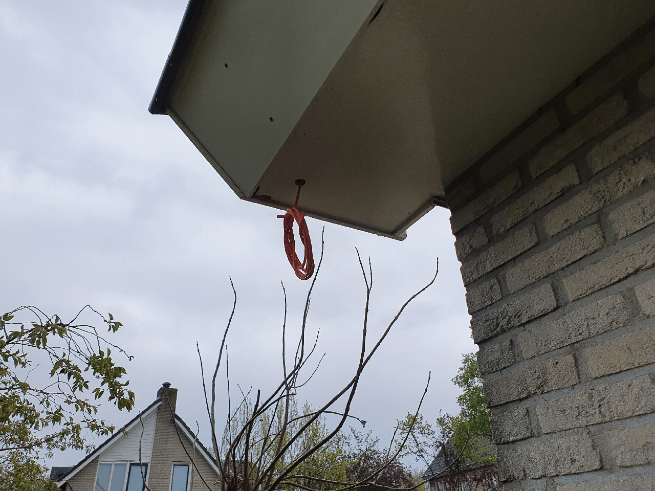 Opgerolde oranje netwerkkabel hangt klaar uit een boorgat in de dakoverstek van de woning, voordat de camera wordt gemonteerd