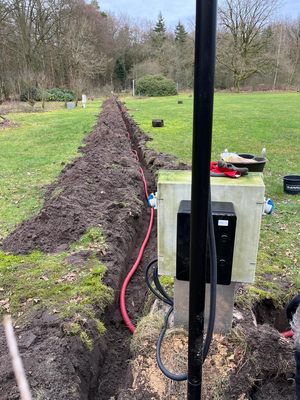 Mast met aansluitkast op het terrein, met daarnaast de open sleuf en de rode mantelbuis die naar de kast toe loopt