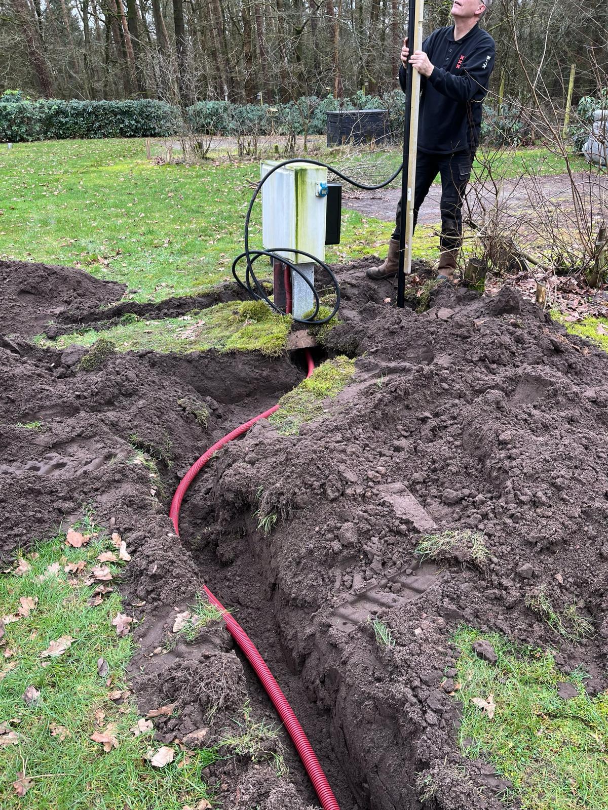 Monteur van CameraInstallatie zet met een stok een mast op zijn plek bij een aansluitkast, met de rode mantelbuis in de sleuf ervoor