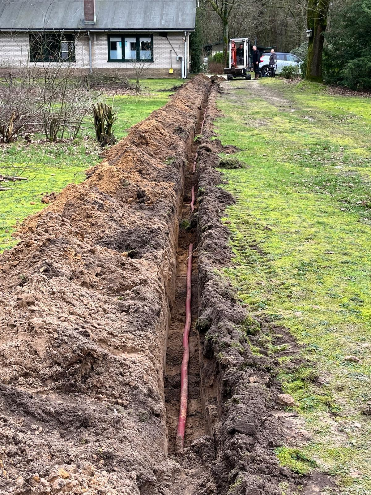 Lange open sleuf dwars over het grasveld van het recreatieterrein met een rode mantelbuis erin, richting een gebouw
