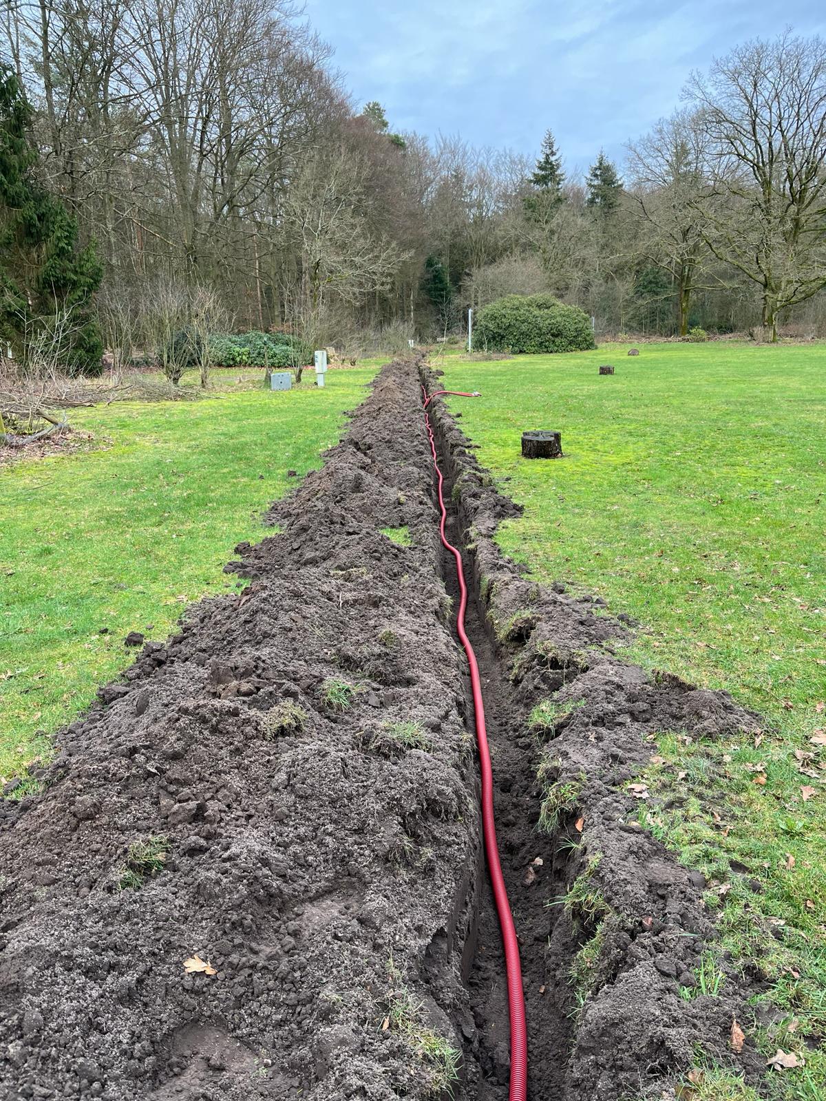 Open sleuf met rode mantelbuis dwars over het grasveld van het recreatieterrein richting de bosrand