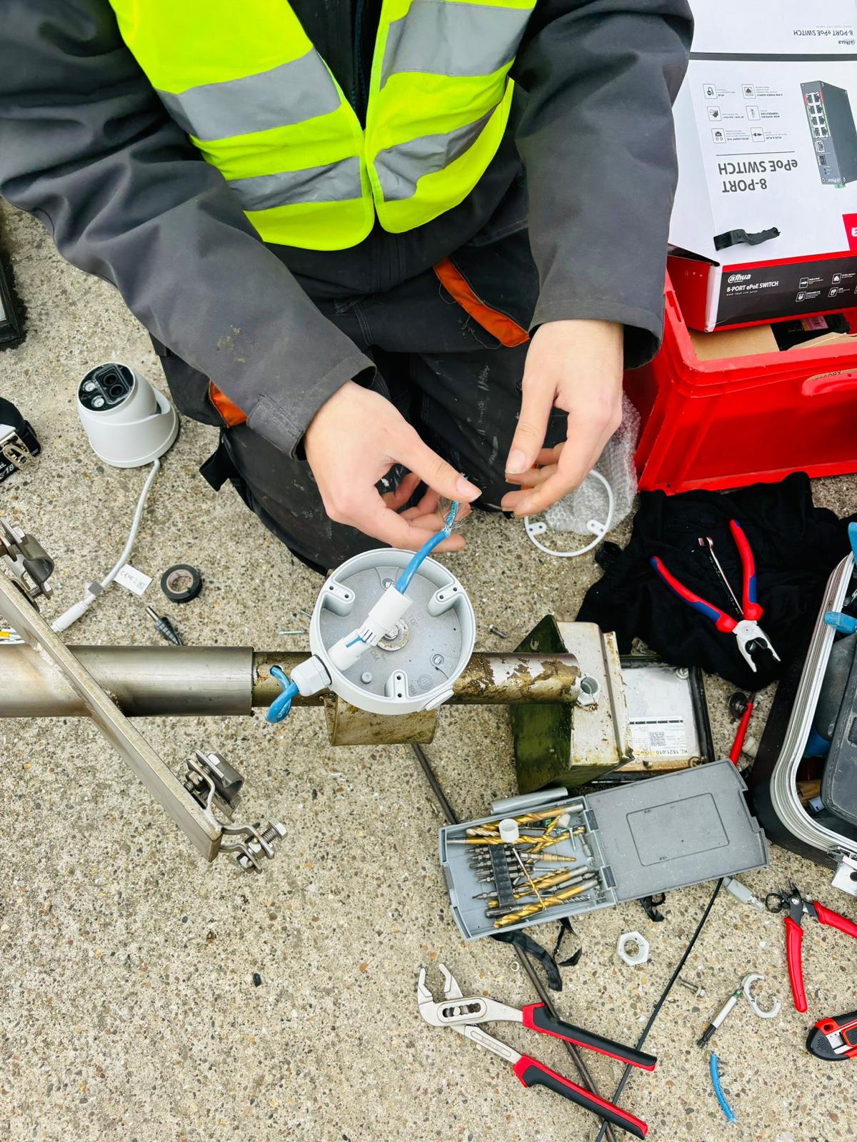 Monteur monteert een aansluitkast met PoE-switch op een mast, met gereedschap en materiaal uitgestald op de grond