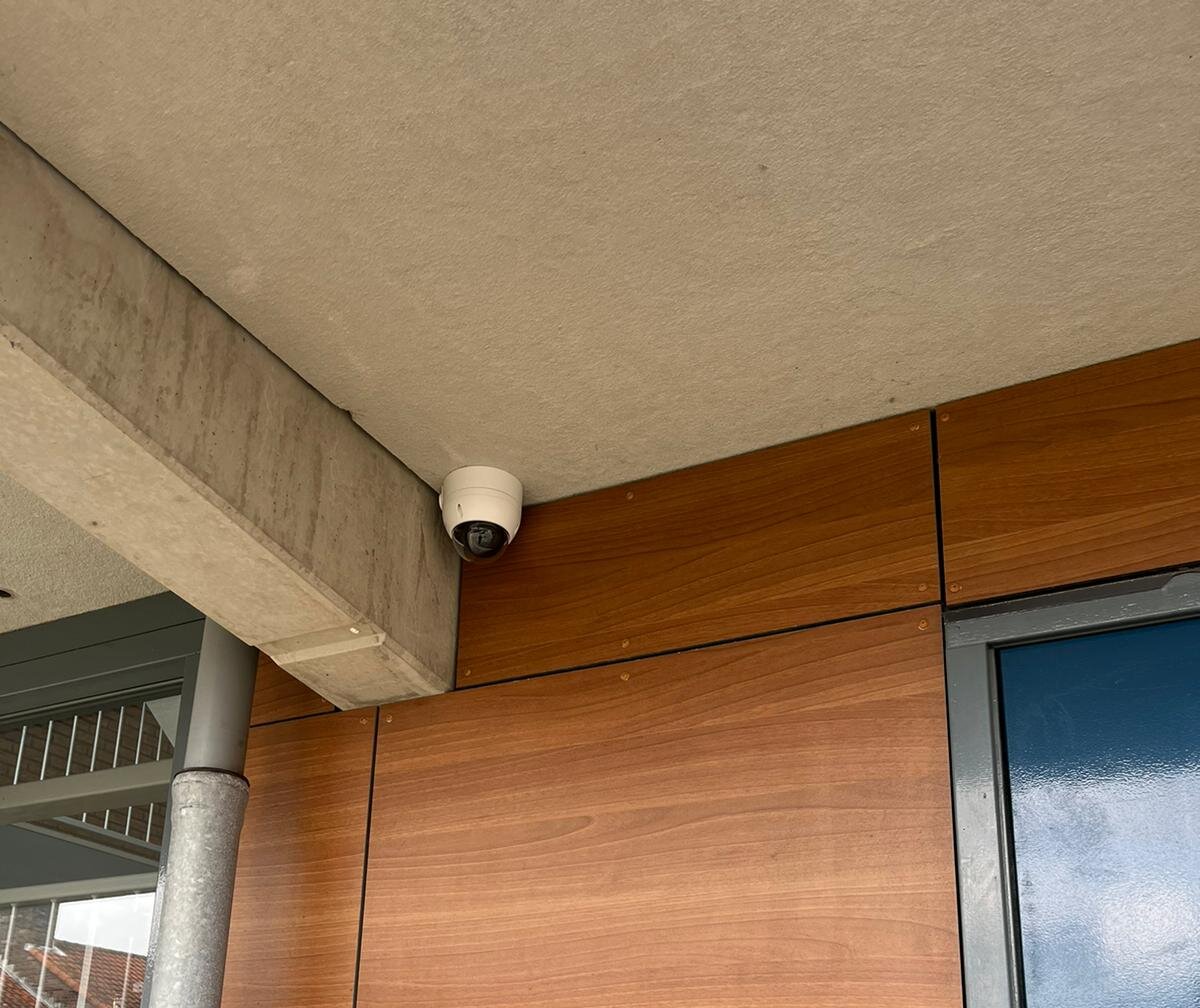 Witte dome-camera onder een betonnen luifel tegen houten gevelbekleding bij de entree van een wooncomplex