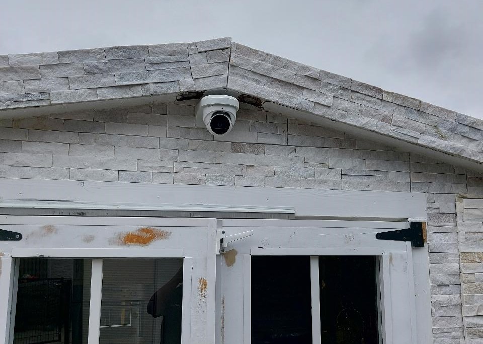 Witte turret-camera onder de puntgevel van een bijgebouw met lichte steenstrips, gericht op de toegangsdeuren eronder