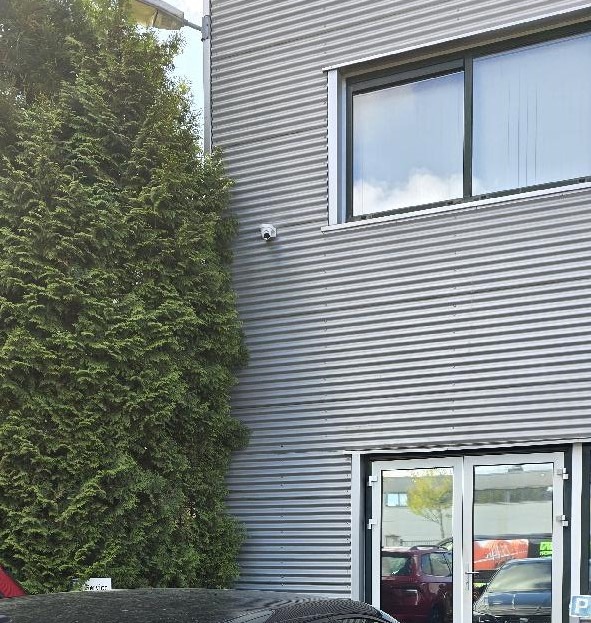Dome-camera op een grijze golfplaten bedrijfsgevel naast een hoge conifeerhaag