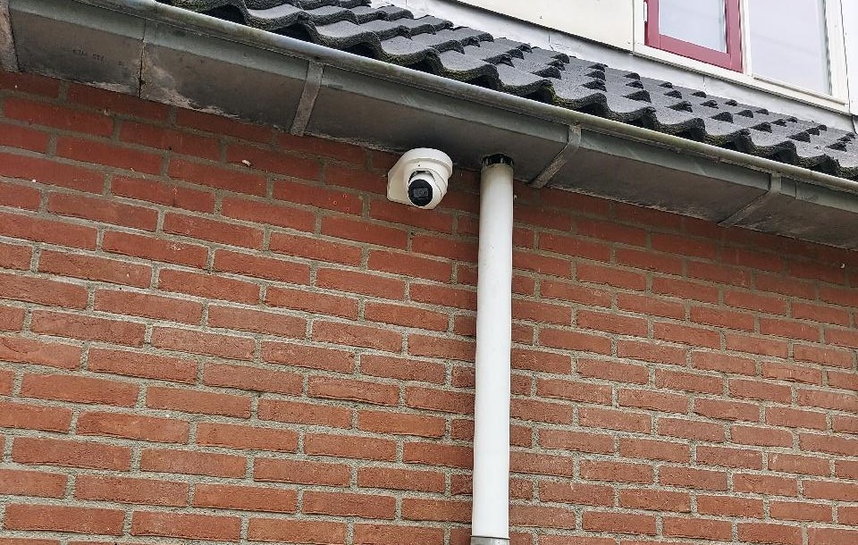 Witte turret-camera strak onder de goot van een woning met rode baksteen, direct naast de regenpijp