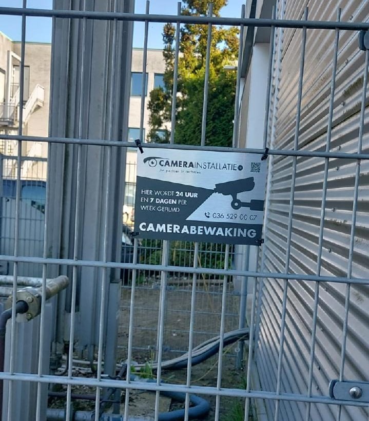 Bord met de tekst camerabewaking van CameraInstallatie bevestigd aan een stalen hekwerk op het terrein
