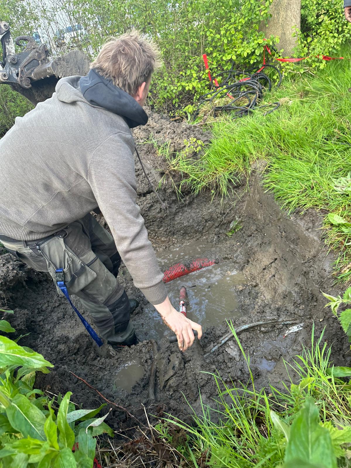 Monteur van CameraInstallatie legt in een natte, modderige sleuf een rode mantelbuis op zijn plek naast de bak van een graafmachine