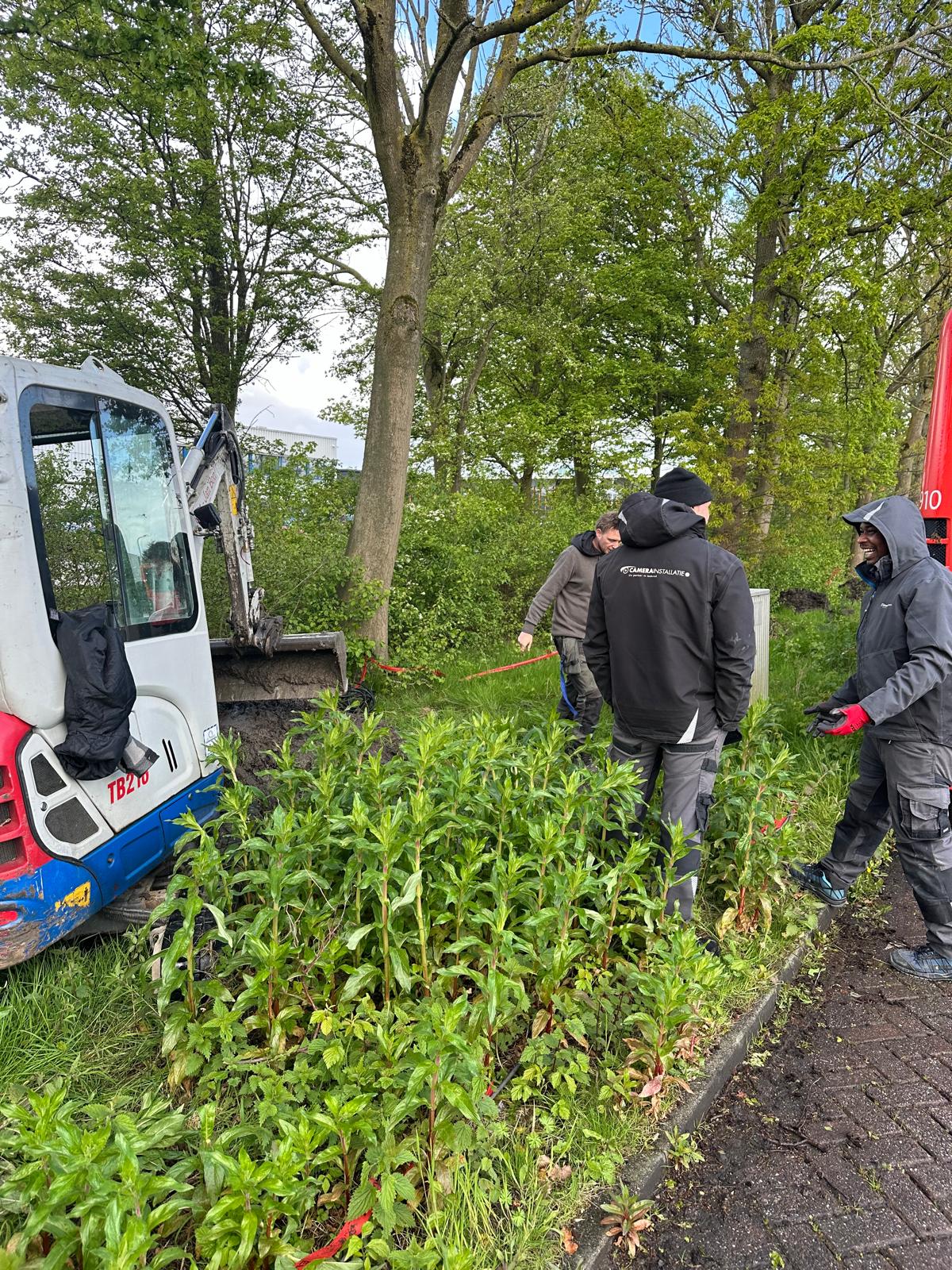 Monteurs van CameraInstallatie overleggen bij een minigraver naast een openliggende sleuf op het terrein