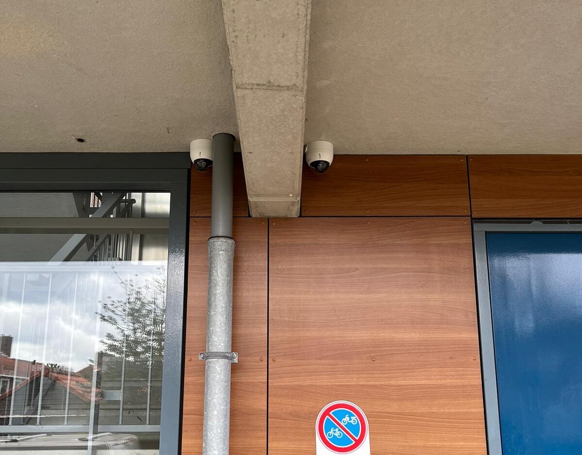 Twee witte dome-camera's onder een betonnen luifel bij de entree van een appartementencomplex met houten gevelbekleding