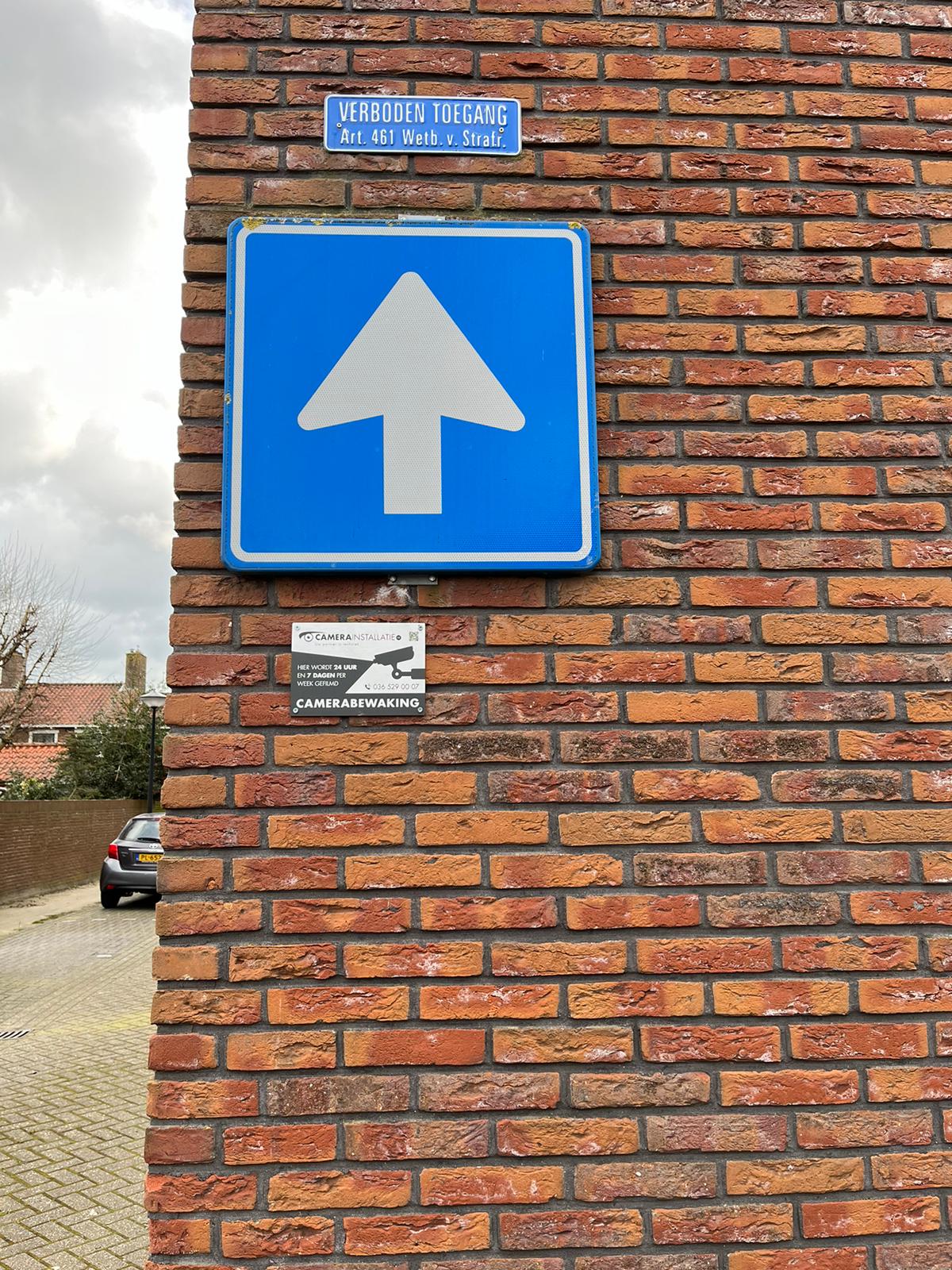 Sticker met de tekst camerabewaking onder een verkeersbord op een bakstenen gevel bij de ingang van een woonterrein