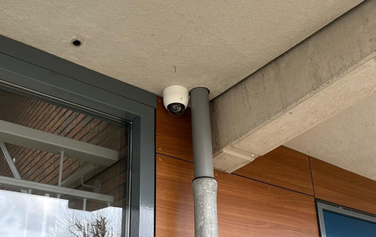 Witte dome-camera onder een betonnen luifel naast de glazen entreedeur van een appartementengebouw