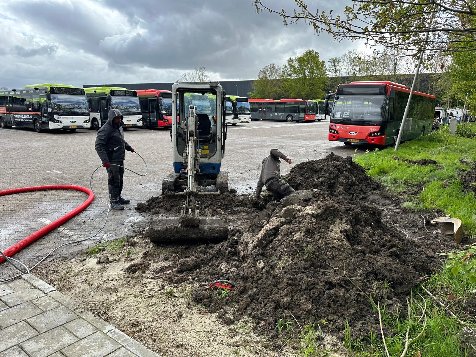 Stallingsterrein met geparkeerde stadsbussen, een minigraver en een open kabelsleuf met rode mantelbuis waarin monteurs van CameraInstallatie werken