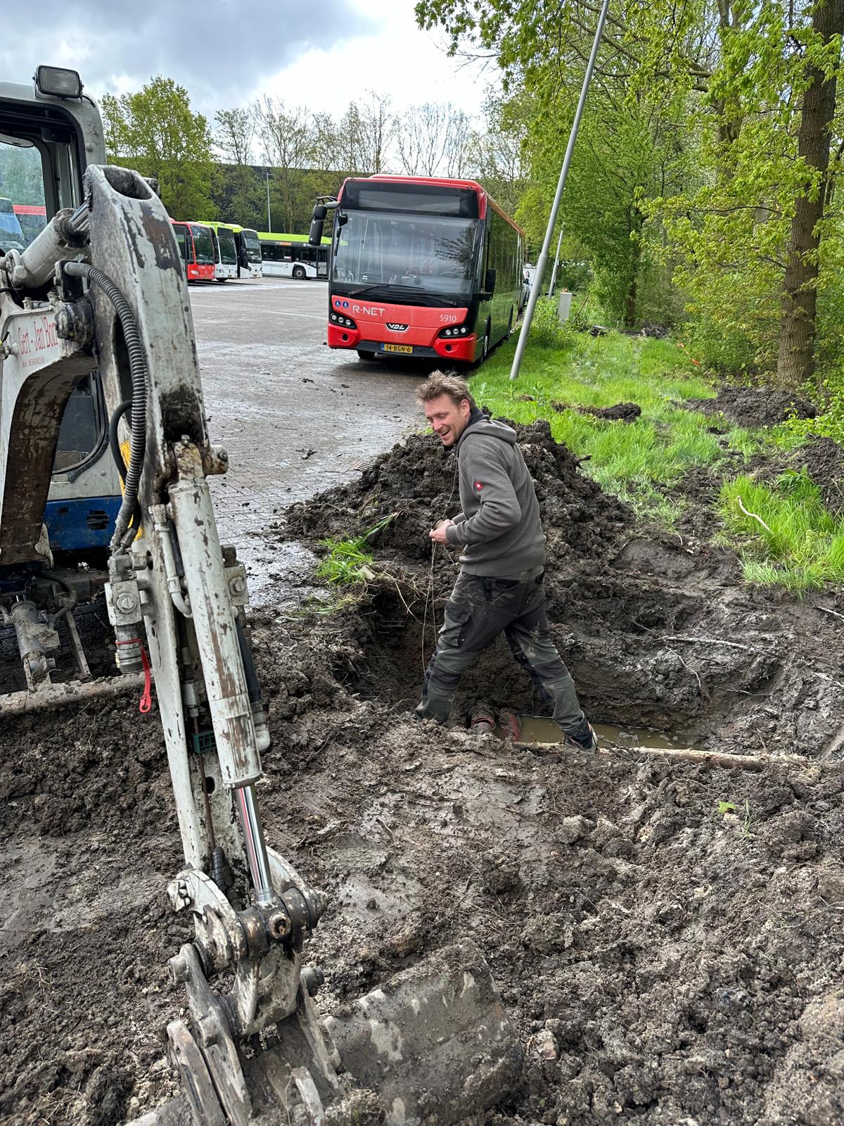 Monteur van CameraInstallatie staat in een verse kabelsleuf op het terrein, met achter hem een geparkeerde stadsbus en een minigraver