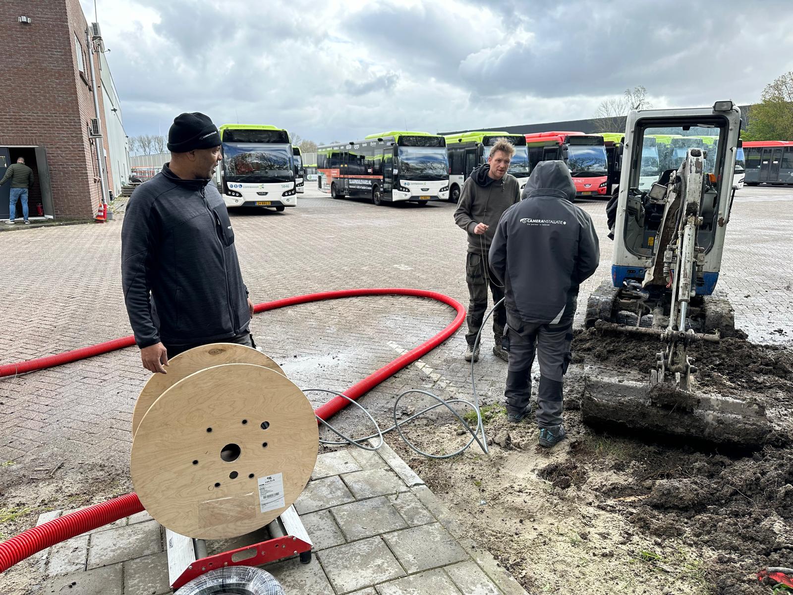 Monteurs van CameraInstallatie bij een houten kabelhaspel en rode mantelbuis naast een minigraver, met op de achtergrond een rij geparkeerde stadsbussen