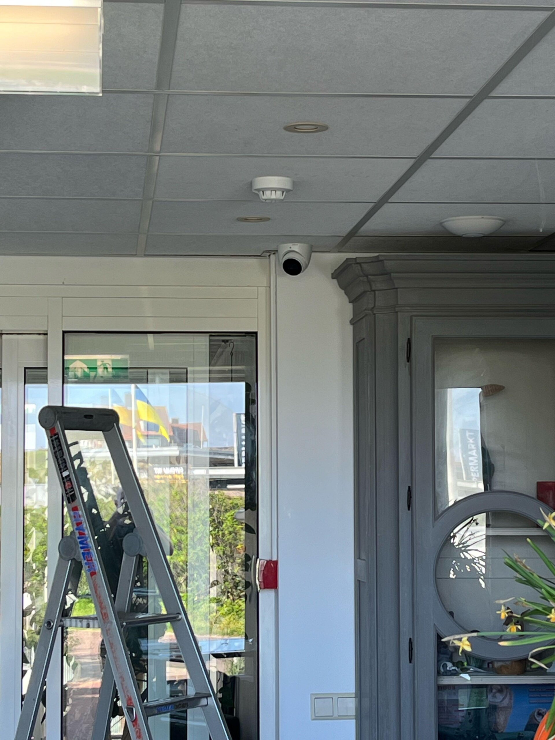 Witte dome-camera binnen bij de entree van het hotel, gemonteerd op de hoek boven de schuifdeur, met een trap van de monteur ervoor