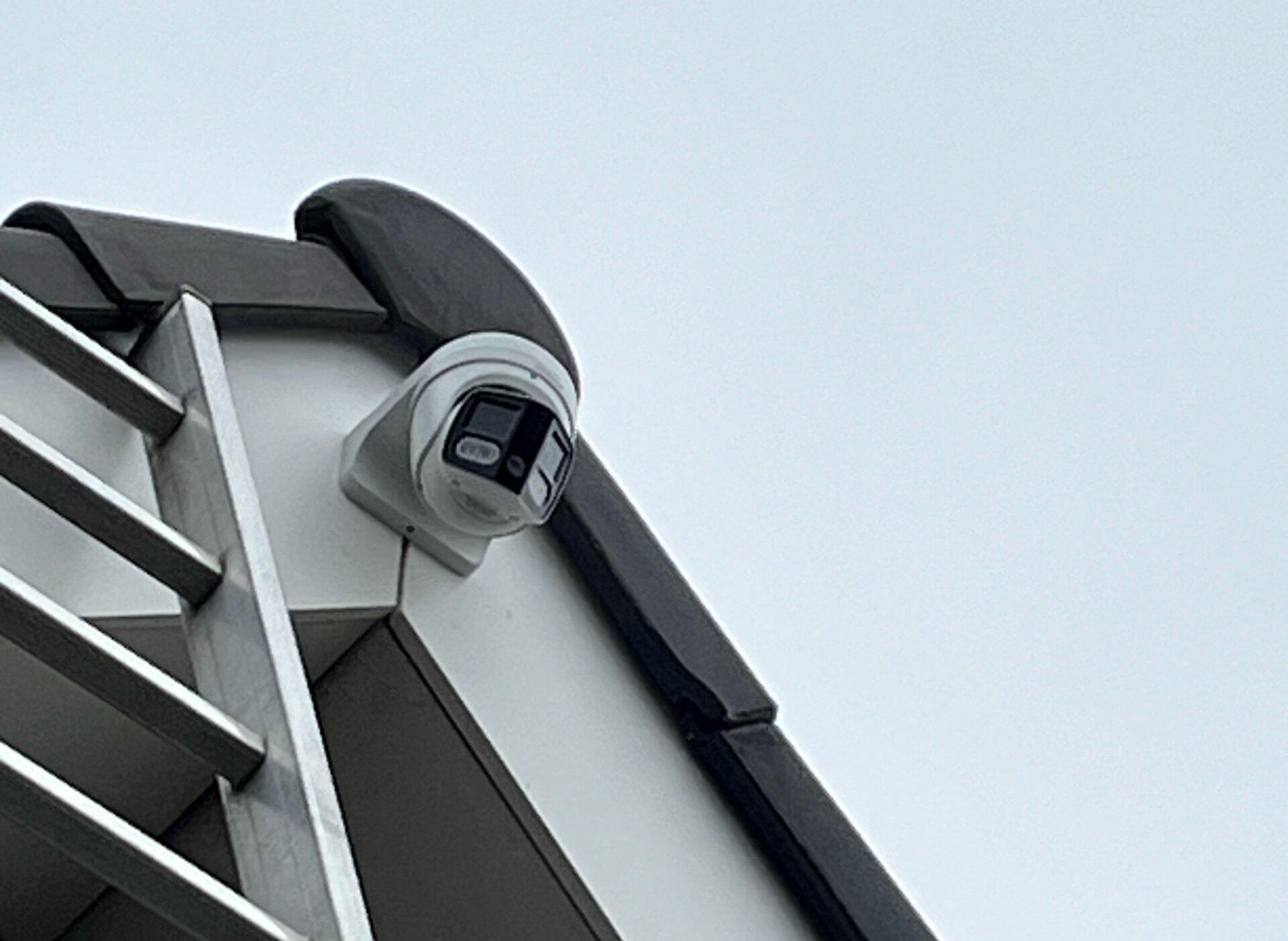 Beveiligingscamera met twee lenzen gemonteerd onder de dakrand van een wit gebouw, met een ladder ertegenaan tijdens de montage