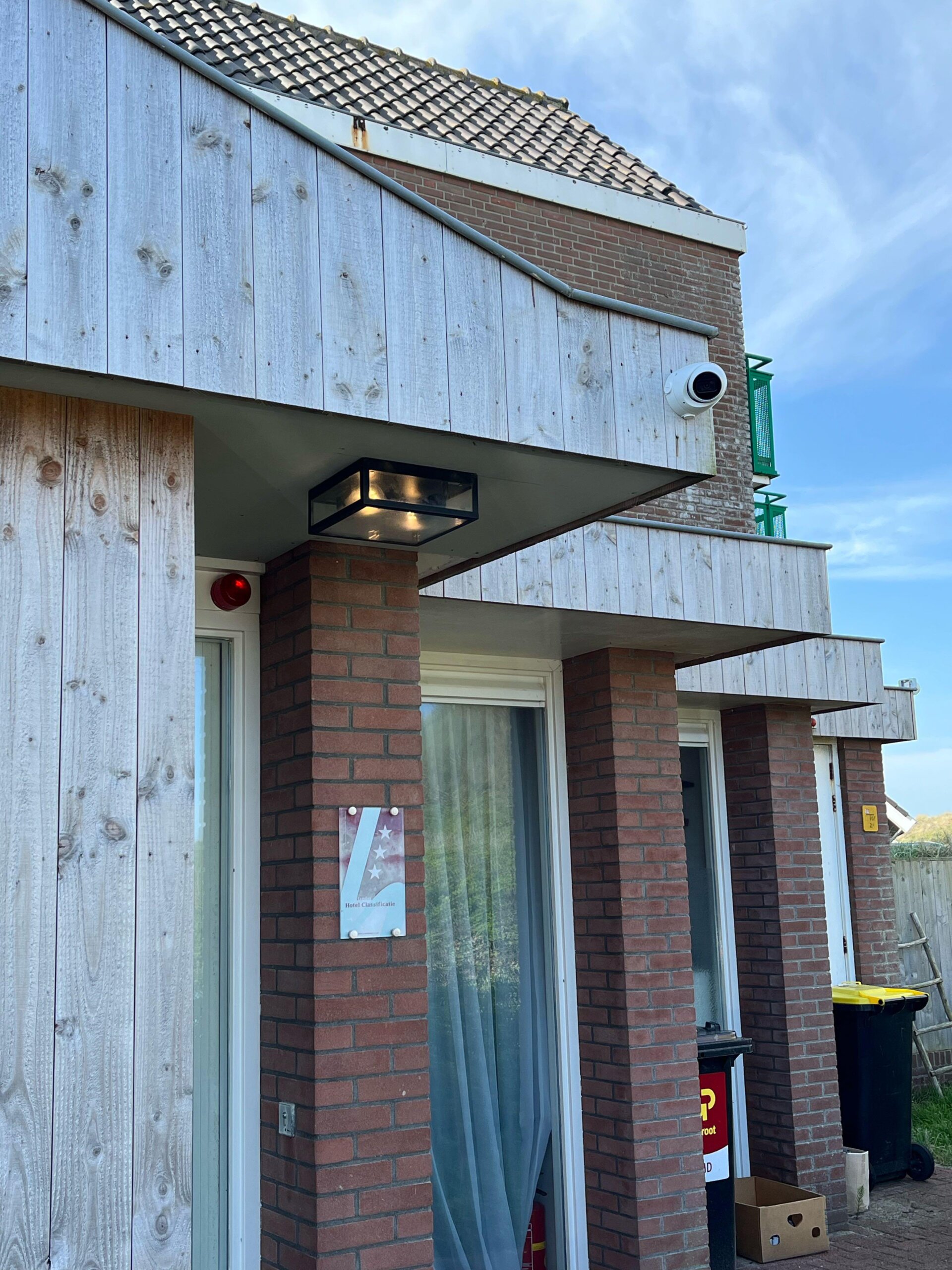 Witte beveiligingscamera onder de houten gevelbetimmering van een hotelgebouw aan zee, naast de entree met een plaquette hotelclassificatie