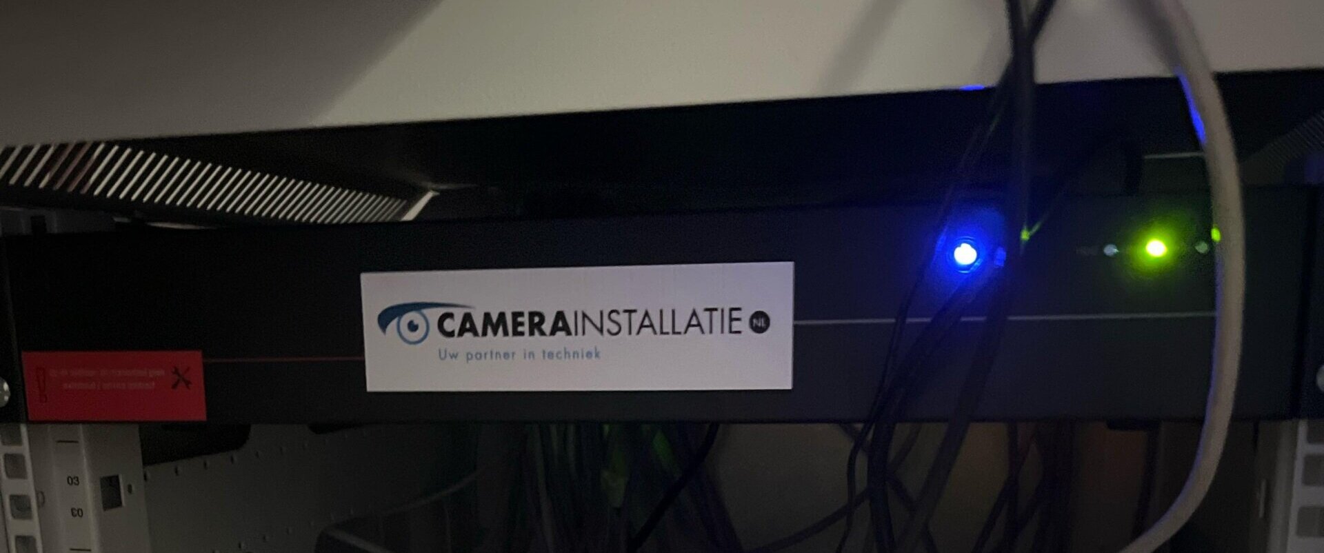 Zwarte apparatuur in een 19-inch rack met brandende blauwe en groene statuslampjes en een sticker van CameraInstallatie