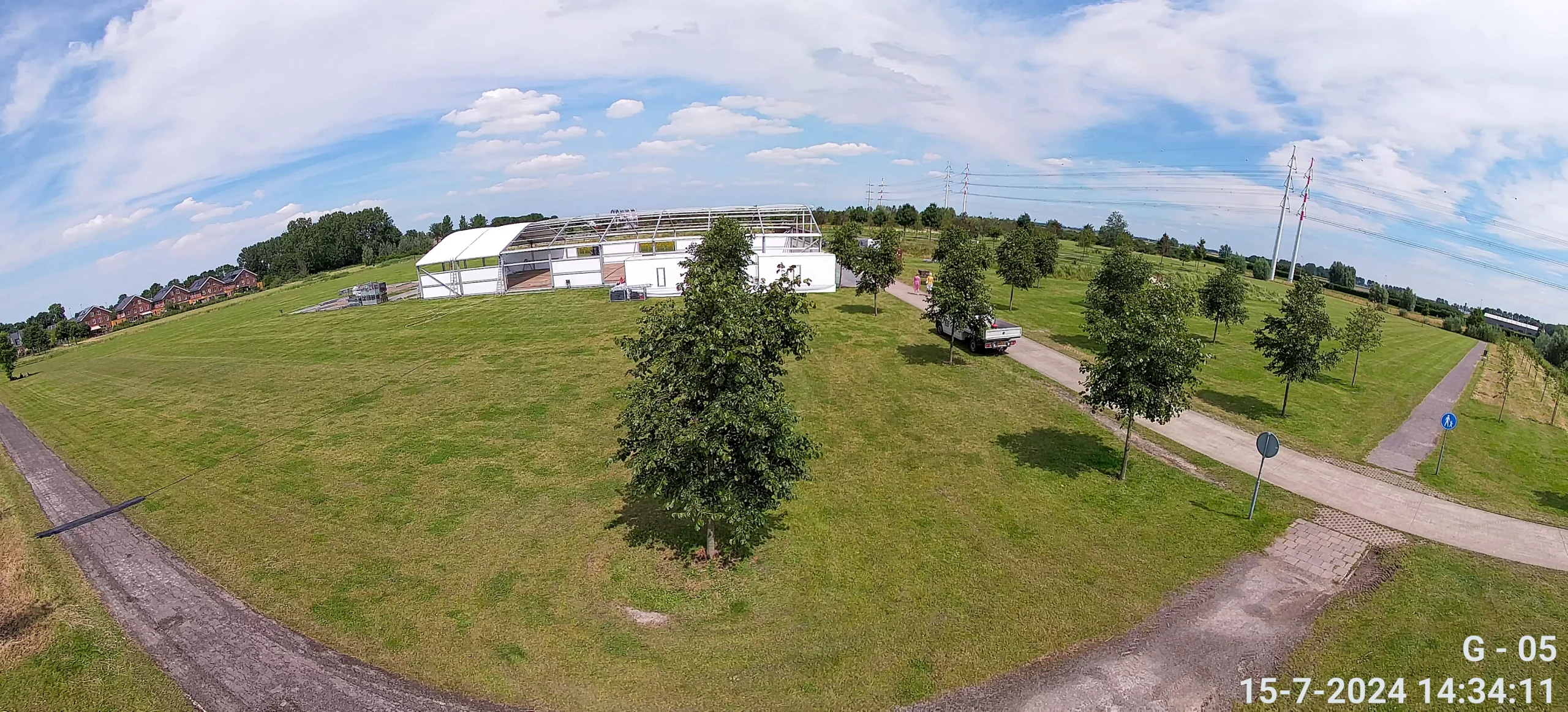 Camerabeeld vanaf een mast: breed overzicht over een grasveld met een grote tent, een pad en bomen