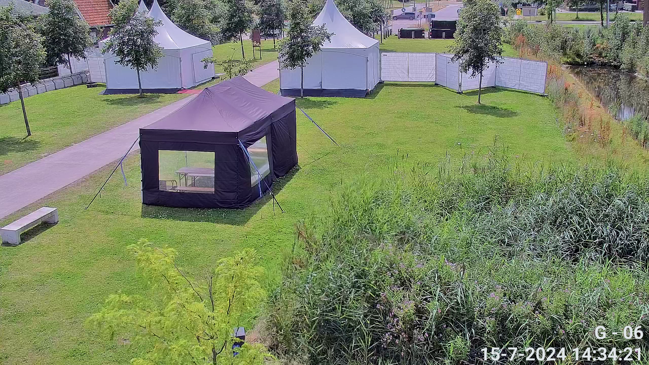 Camerabeeld vanaf een mast op een grasveld met partytenten, hekwerk en een sloot
