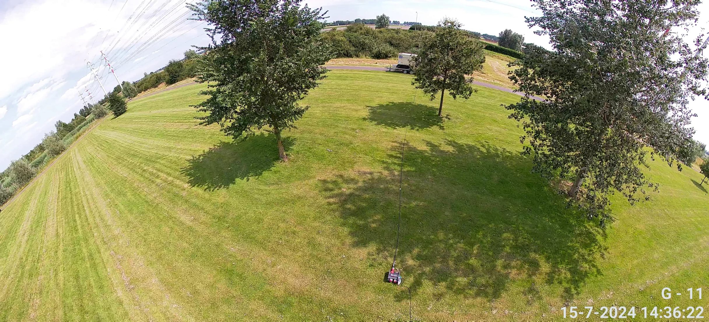 Camerabeeld vanaf een mast over een leeg, pas gemaaid grasveld met bomen en een toegangsweg op de achtergrond