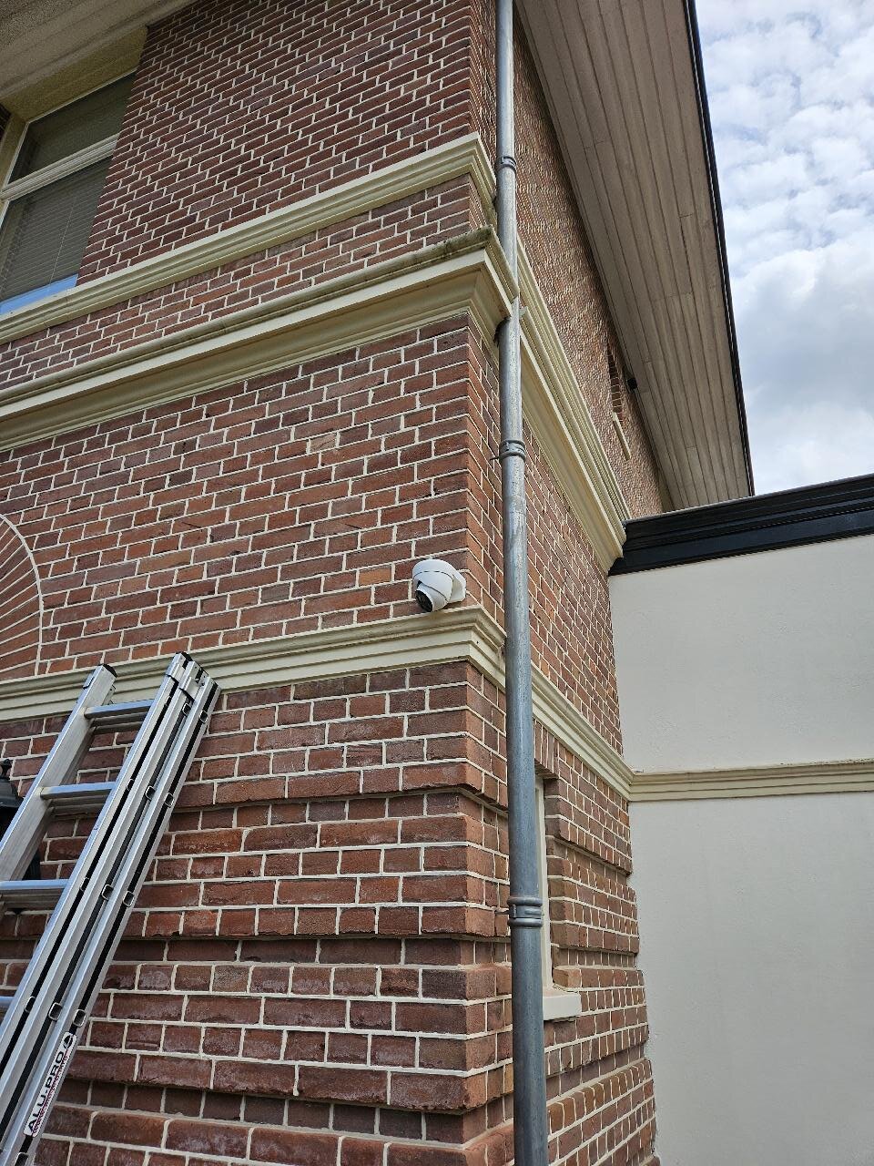 Witte dome-camera op de hoek van een bakstenen gevel, gemonteerd vlak boven de sierlijst naast de regenpijp
